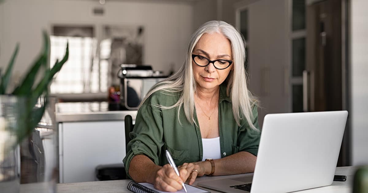 Senior stylish woman taking notes in notebook while using laptop at home. Old freelancer writing details on book while working on laptop in living room. Focused cool lady writing notary in notepad.